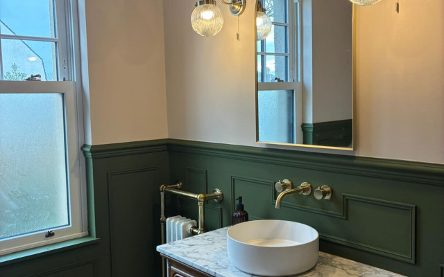 A modern bathroom showcasing green walls complemented by gold trim details, featuring elegant lighting and a stylish wooden vanity.