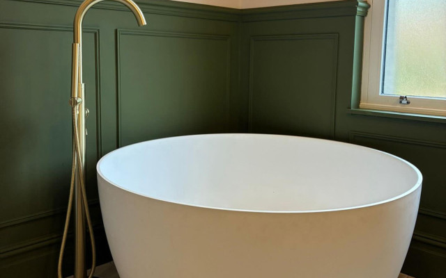 A white bathtub stands elegantly in a bathroom with deep green walls, complemented by a stylish gold tap, creating a fresh and inviting atmosphere.