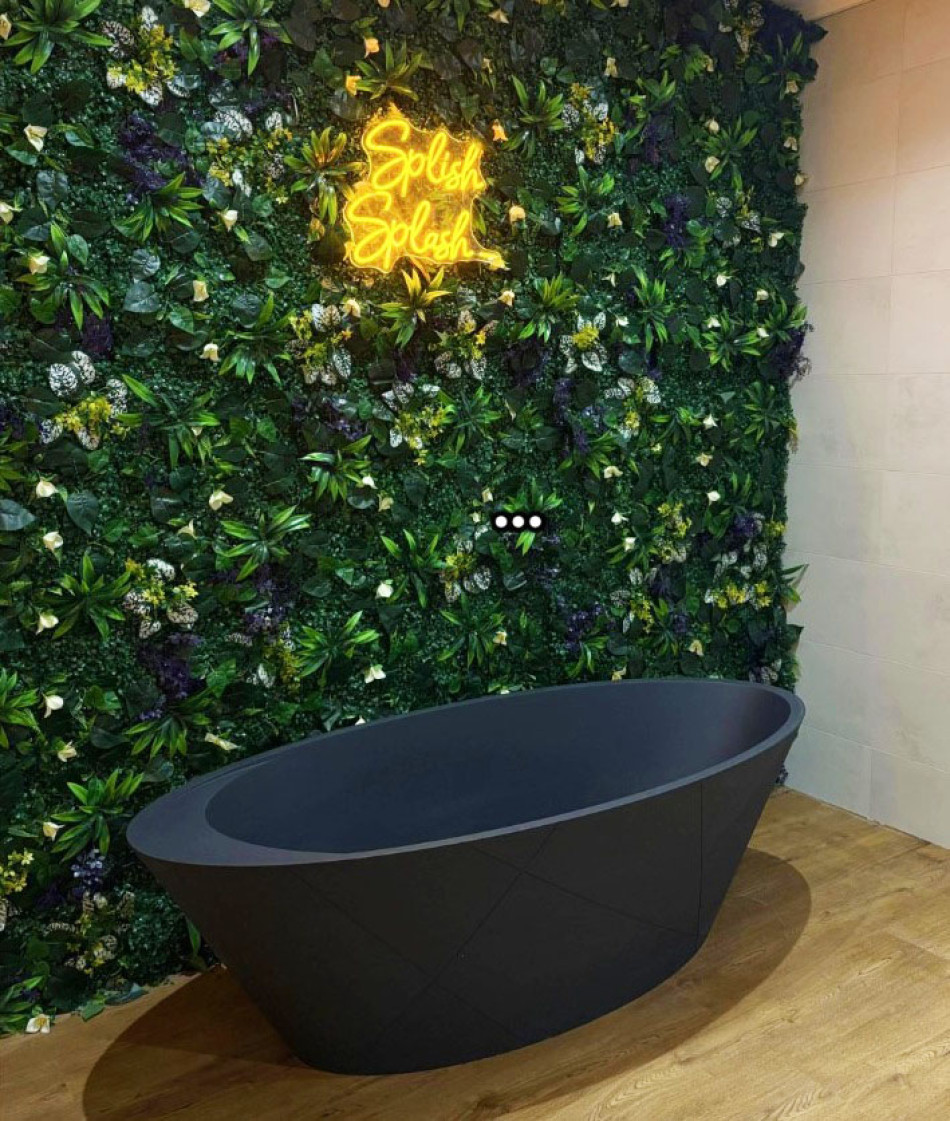 A modern bathroom showcasing a sleek black bathtub set against a vibrant green wall, accented by a glowing neon sign reading 'Splash Splash'.